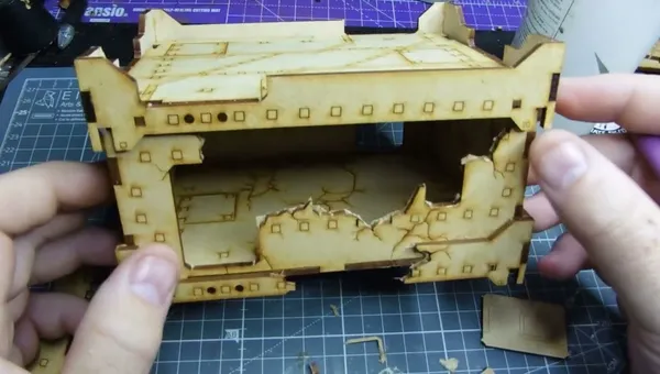 A partially assembled, light brown cardboard or MDF terrain piece with a damaged, jagged top edge, revealing an open interior. The structure has square and rectangular cutouts, with small square and circular holes along the edges. It rests on a green self-healing cutting mat with a grid pattern. A hand is visible on the left side, holding part of the terrain. Small debris and tools, including a metal L-shaped piece, are scattered on the mat.