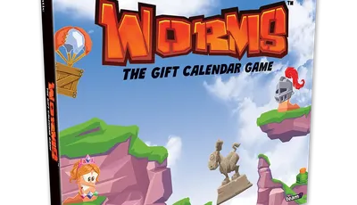 A colorful box for "Worms: The Gift Calendar Game" features a vibrant illustration of cartoonish landscapes with green hills and rocky cliffs. Prominent characters include a pink worm wearing a crown and a knight, both positioned on the cliffs, while a hot air balloon floats in the sky. The title is boldly displayed at the top, emphasizing the festive theme of the advent calendar.