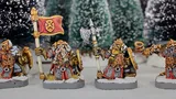 Three painted miniature figures of fantasy dwarves stand on round bases in a snowy outdoor setting with blurred evergreen trees in the background.

The left figure wears a horned helmet, patterned tunic, and fur cloak, holding a pole-mounted red banner with a gold emblem of a snarling face. The figure also carries a round shield on their back.

The center figure, slightly behind the others, wears a helmet with a nose guard, a fur-lined cloak, and carries a pole weapon with a curved blade.

The right figure, holding a large double-bladed axe over one shoulder, wears a horned helmet, fur cloak, and patterned tunic. Their beard is braided and wrapped around their waist.