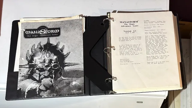A three-ring binder containing the playtest manuscript for "ManaStorm: The Magic Adventure Game," featuring a black and white cover illustration of a character with a spiked helmet and a weapon. The title "MANASTORM The Magic Adventure Game" is prominently displayed at the top, along with the version number and date on the first page inside the binder. Additional pages include loose character sheets and player handouts, indicating the game's mechanics and spells.