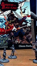 A vibrant tabletop scene features the rulebook for "Boarders and Black Flags" by Man ’o’ Kent Games, prominently displayed in the center. The cover art depicts a dynamic pirate battle on a ship, with characters wielding swords and engaging in combat. Surrounding the book are several unpainted miniatures, including human pirates and undead Skelliwags, set against a backdrop of MDF scenery elements like masts and crates, all arranged on a wooden table.