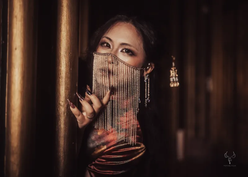A woman with long, dark hair poses against a wooden pillar, partially obscured by a decorative chain veil that hangs over her face. She wears a rich, dark-colored dress with floral patterns and has long, pointed nails, adding to her dramatic appearance. The background features warm, muted tones, enhancing the gothic atmosphere suitable for a vampire-themed event.