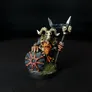 A painted miniature figure of a Chaos Dwarf warrior with a long orange beard, wearing dark armor and a helmet with large curved horns. The figure holds a round shield with a red star design and a spear topped with a skull. The base has small patches of green grass-like texture. The background is black.