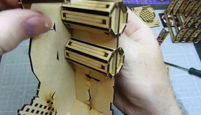 A close-up view of a partially assembled Orc Mega Bunker model from TT Combat, specifically the Orc Sektor section. The image shows a hand holding a piece of the model, which features intricate cut-out designs and layered components. The background includes a grid pattern, suggesting a crafting or modeling workspace.