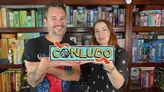 Two people holding a sign reading "CONLUDO" with board games on shelves behind them.