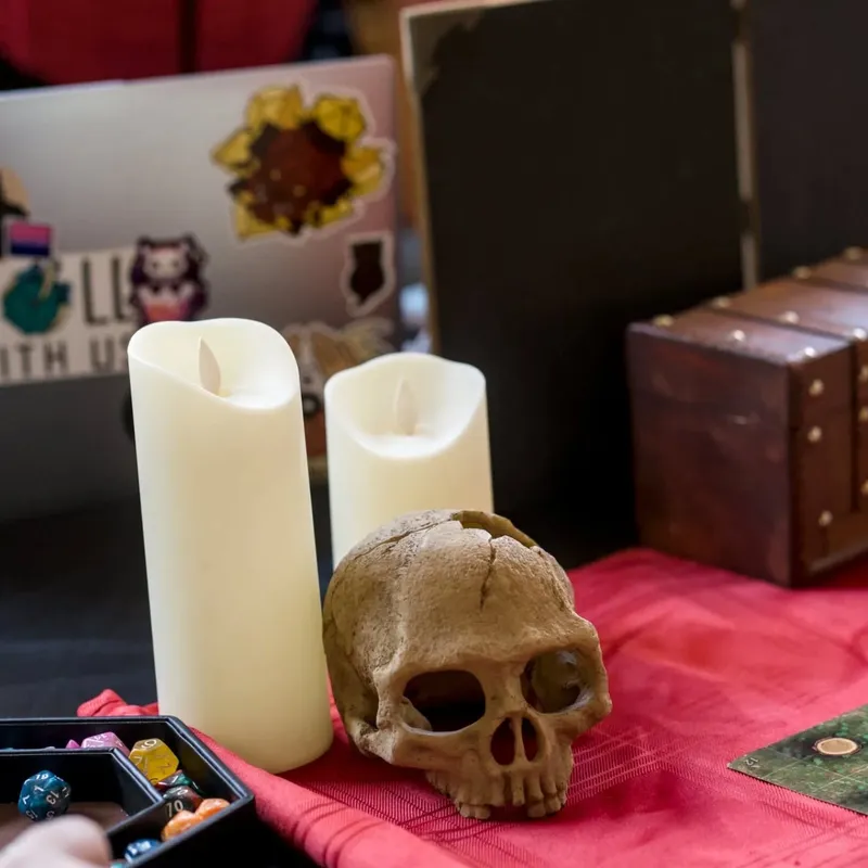 A tabletop scene features two white candles and a decorative skull, set against a red cloth. Nearby, a black tray holds various polyhedral dice, while a small wooden chest is partially visible in the background. The overall arrangement evokes a gothic and mysterious atmosphere, fitting for a vampire-themed gaming event.