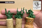 Three green goblin shaman miniatures are displayed on top of cork bases, showcasing their intricate details. The goblins feature various accessories, including staffs and bags, with distinct facial expressions that convey mischief. The background is a neutral color, allowing the vibrant green of the miniatures to stand out prominently.