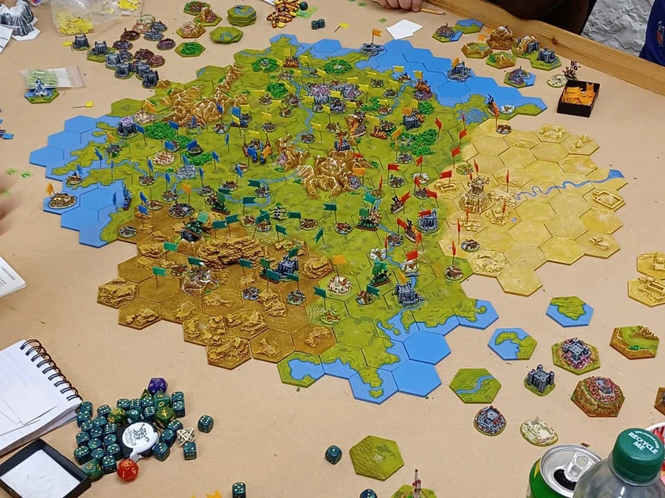 A detailed tabletop gaming scene features a large hexagonal map covered in various terrain types, including green fields, golden hills, and blue water. Numerous miniature figures are positioned across the map, with flags indicating different territories, and several dice are scattered nearby. In the foreground, a notepad and a collection of dice in various colors are visible, suggesting active gameplay.