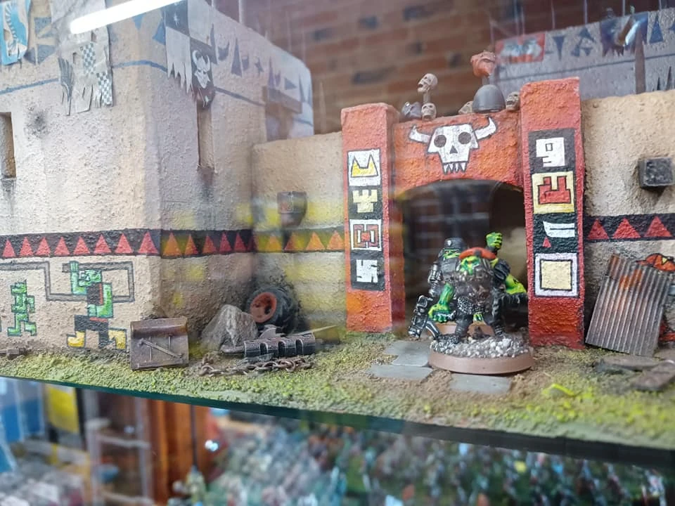 A detailed miniature display features a vibrant, stylized entrance with a skull motif and colorful tribal patterns, set against a textured wall. In the foreground, two miniatures are positioned: one is a green-skinned character with a red shoulder pad, and the other is a darker figure, both appearing ready for action. Surrounding them are various terrain elements, including rocks, a tire, and a wooden crate, enhancing the scene's immersive quality.