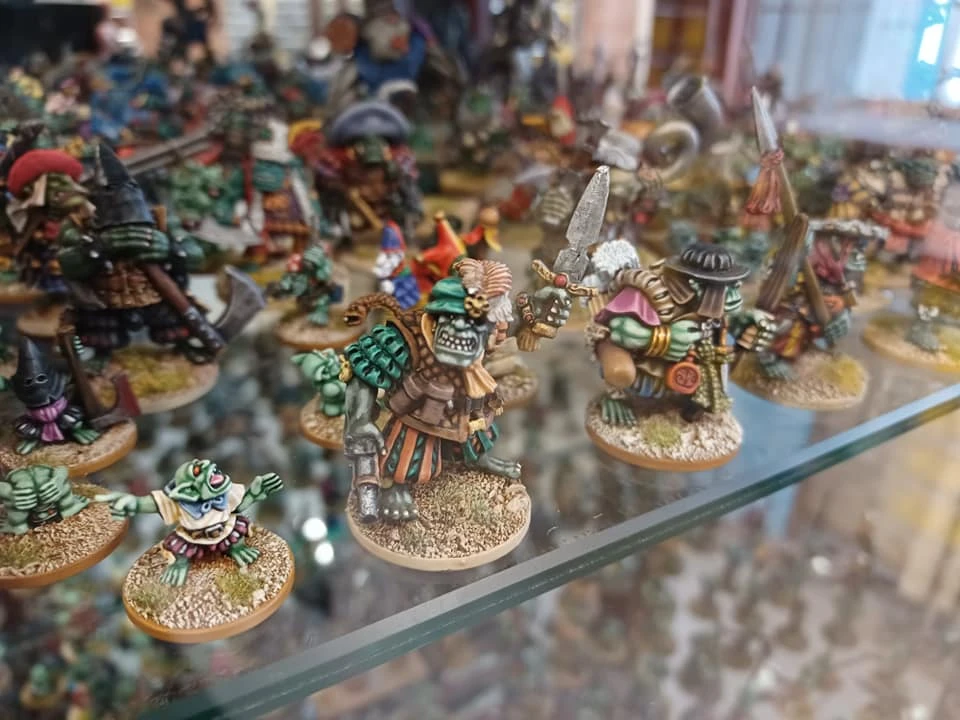 A display case filled with a variety of painted miniature figures, primarily featuring green-skinned goblins and orcs. The foreground showcases a larger orc wearing armor and holding a sword, alongside smaller goblins in playful poses. The background is populated with more miniatures, including various fantasy-themed characters and creatures, all set against a textured base that mimics a natural terrain.