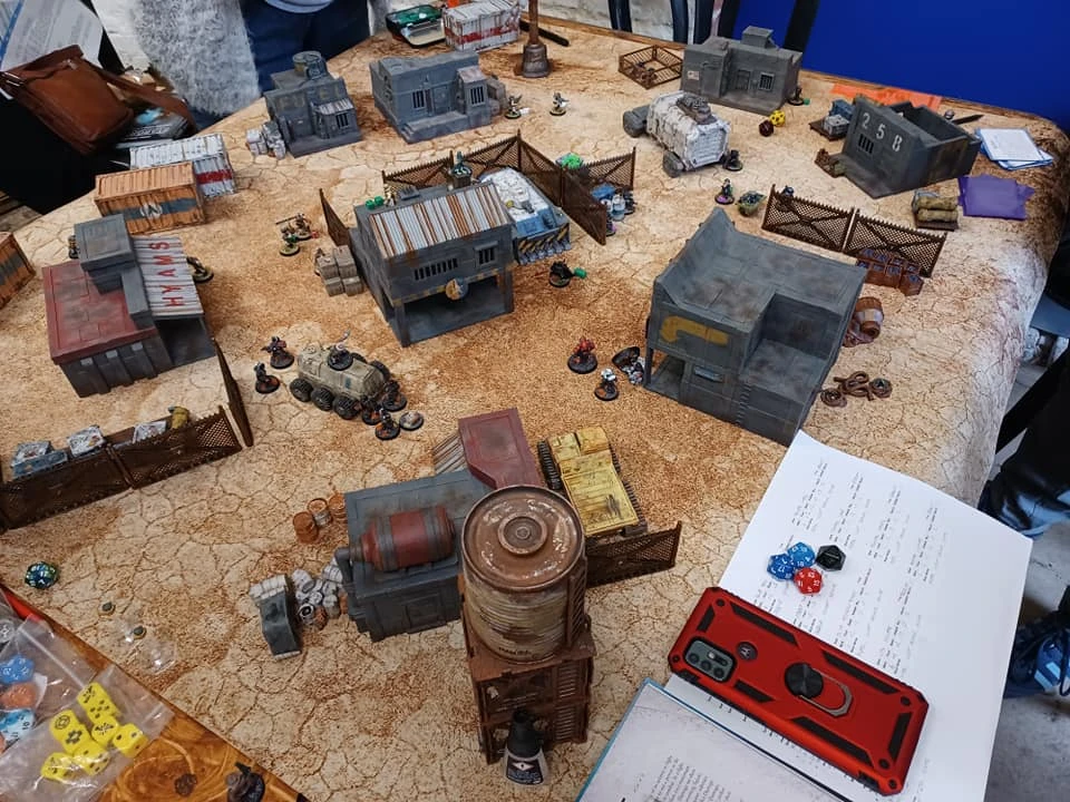A tabletop gaming scene features a detailed battlefield with various terrain elements, including buildings, fences, and scattered debris. Miniature figures are positioned throughout the area, suggesting an active game in progress. In the foreground, a notebook and a set of colorful dice are visible, alongside a smartphone, indicating players are keeping track of game mechanics.