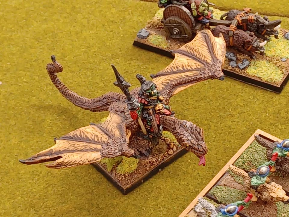 A detailed miniature scene features a dragon with outstretched wings, carrying a rider adorned in colorful armor and wielding a spear. In the background, a cart pulled by two boar-like creatures is visible, along with a grassy terrain dotted with small patches of foliage. The overall setting evokes a classic Oldhammer aesthetic, showcasing the vibrant colors and intricate details typical of tabletop gaming miniatures.