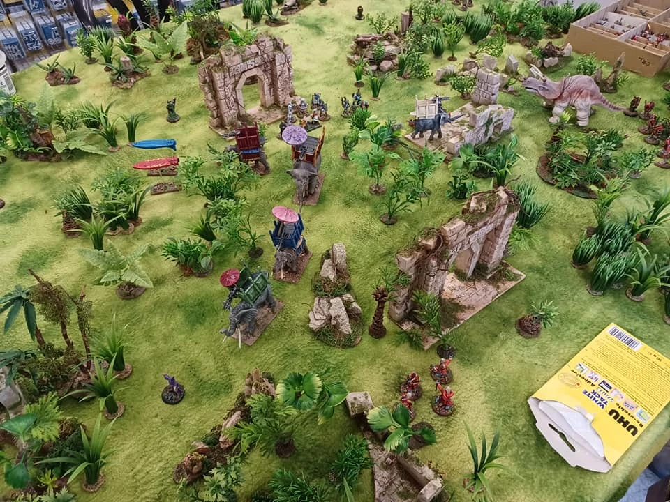 A tabletop gaming scene features a lush green terrain scattered with various miniature figures and structures. In the center, there are ruins made of stone, surrounded by dense foliage and palm-like plants, while several colorful banners and structures, including small towers and elephants, add to the vibrant atmosphere. A few dinosaur figures are also visible, contributing to the fantastical setting of the game.