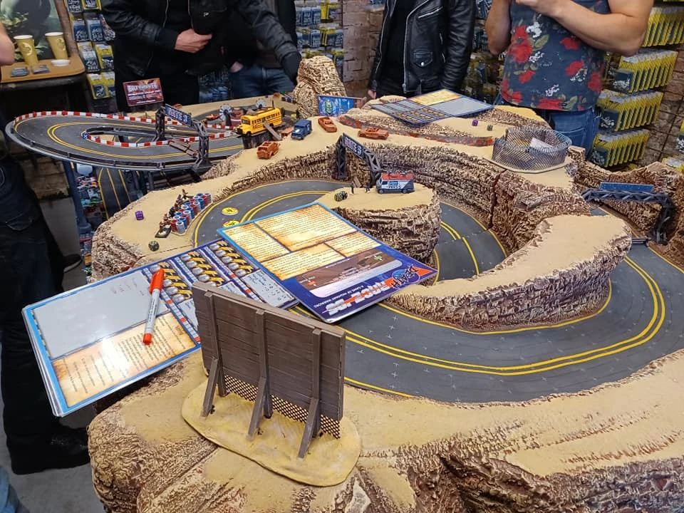 A detailed tabletop gaming setup features a winding race track made of textured terrain, resembling rocky cliffs and sandy surfaces. Miniature vehicles are positioned along the track, with various game components, including dice and rulebooks, scattered on the table. In the background, several people are engaged in the game, adding to the lively atmosphere of the event.