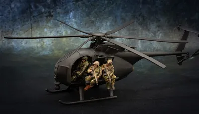 A detailed model of a Little Bird helicopter, featuring a dark color scheme, is prominently displayed. Two miniature soldiers are seated on the helicopter's skids, equipped with rifles, while another soldier is visible inside the cockpit. The background features a textured gradient, enhancing the focus on the helicopter and its crew.