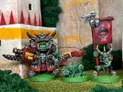 A trio of space orcs stands prominently in the foreground, featuring Attila the Warlord, who is adorned in elaborate armor with spikes and a large horned helmet, wielding a futuristic weapon. Beside him is a small green orc hound, Snaarl, and Groblett the Goblin, who holds a banner displaying a skull and horns. The background includes stylized stone structures and greenery, enhancing the fantastical setting.
