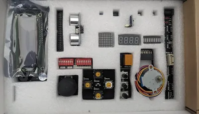 A collection of electronic components is neatly arranged on a white foam background. Key items include a pair of ultrasonic sensors, a digital display, various buttons, a relay, and a DIP switch. Additional components like a rotary encoder and several connectors are also present, showcasing a variety of parts typically used in DIY electronics projects.