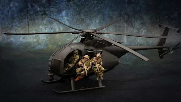 A detailed model of a Little Bird helicopter, featuring a dark color scheme, is prominently displayed. Two miniature soldiers are seated on the helicopter's skids, equipped with rifles, while another soldier is visible inside the cockpit. The background features a textured gradient, enhancing the focus on the helicopter and its crew.
