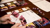 A tabletop gaming scene features a wooden table with various gaming materials. Prominently displayed is the "Daggerheart Class Pack" for the Bard, showcasing vibrant artwork on its cover. Nearby, a player holds a stack of character cards, while several polyhedral dice and character sheets are scattered across the table, indicating an active game session.