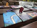 A red wooden game piece standing on a wooden board game tile with a blue and green landscape illustration. Surrounding tiles show varied terrain and paths. Multiple colorful cards with Japanese-inspired artwork and symbols are scattered nearby.