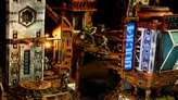 Miniature sci-fi wargame scene with industrial terrain. Three green alien-like figures in combat poses on metal platforms, ladders, and catwalks. Background includes rusted metal structures, exposed pipes, and a glowing blue digital screen displaying binary-like data.