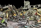 A dynamic scene features a group of detailed miniatures engaged in battle within a ruined, multi-level terrain setting. The backdrop includes crumbling stone walls, wooden scaffolding, and flickering flames, creating a dramatic atmosphere. Among the miniatures are various characters, including a humanoid warrior, a cloaked figure, and a monstrous creature, all poised for action against a new threat.