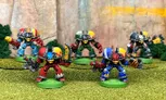 A group of five colorful, 28mm scale space orc miniatures stands on a grassy terrain, each equipped with various weapons and wearing distinctive armor. The orcs are painted in vibrant colors, including red, blue, and green, with unique designs on their helmets and armor, showcasing their cybernetic enhancements. They are posed dynamically, suggesting readiness for battle, against a backdrop of rocky terrain and greenery.
