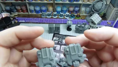 Two gray miniature components are held in each hand, showcasing intricate details such as ridges and small protrusions. In the background, various other gray miniatures and model parts are arranged on a crafting mat, with a selection of paint bottles visible on shelves above. A printed sheet with diagrams and instructions is partially visible beneath the components.