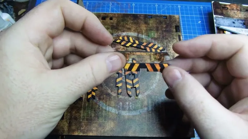 Two hands are shown holding small, printed pieces featuring bold black and orange striped patterns, resembling hazard tape or warning markers. These pieces are being examined against a textured, multicolored background that appears to be a gaming terrain or model base, with various details like grids and circular markings. The workspace is organized with a blue cutting mat underneath, indicating a crafting or modeling activity.