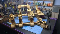 A detailed assembly of the TT Combat Sector 4 Plasma Relay is displayed, featuring intricate wooden structures with multiple towers and connecting pathways. The design includes cylindrical towers with circular tops and various platforms, all set against a blue cutting mat. In the background, jars of paint and tools are visible, indicating a workspace dedicated to tabletop gaming and model building.