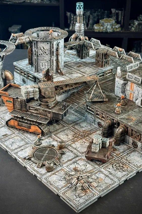 A detailed modular sci-fi terrain setup features a large, industrial-looking vehicle with tracks and a raised platform, surrounded by various structures and pipes. In the scene, two miniature figures are positioned on the terrain, one near a stack of crates and the other on a walkway. The ground is textured with cracked tiles and a circular grate, enhancing the gritty, futuristic atmosphere of the setting.