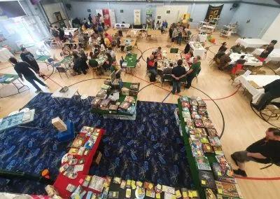 A spacious indoor venue filled with tables where numerous people are engaged in various tabletop games. In the foreground, several tables display a wide array of board games, with colorful boxes and game components visible. The background features groups of attendees playing games, chatting, and enjoying the event, with banners and signs indicating the convention's activities.