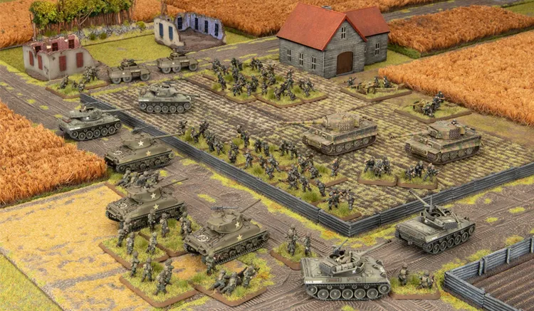 A detailed tabletop wargaming scene depicts a battlefield layout featuring American and German forces from the Flames Of War starter set, Fortress Europe. In the foreground, several M4 Sherman tanks and infantry figures are positioned, while in the background, a mix of German Panzergrenadiers and tanks, including Tiger tanks, are arranged near a small building with a red roof. The terrain includes fields, roads, and scattered structures, creating a dynamic and immersive environment for gameplay.