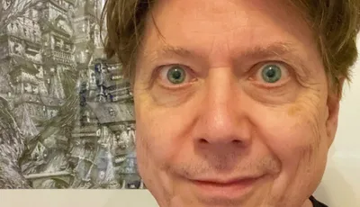 A close-up portrait of a man with light brown hair and striking green eyes, smiling slightly. He is wearing a black shirt and is positioned in front of a detailed, artistic background featuring a complex, fantastical structure, possibly a building or landscape. The overall tone of the image is casual and inviting, with a focus on the subject's expressive features.