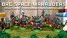 A group of five intricately detailed space orc miniatures stands on a grassy base, showcasing their vibrant colors and unique designs. Each orc is heavily armed, with some wielding axes and others holding futuristic weapons, adorned with cybernetic enhancements and horned helmets. The backdrop features a textured stone wall, enhancing the sci-fi theme of the scene.