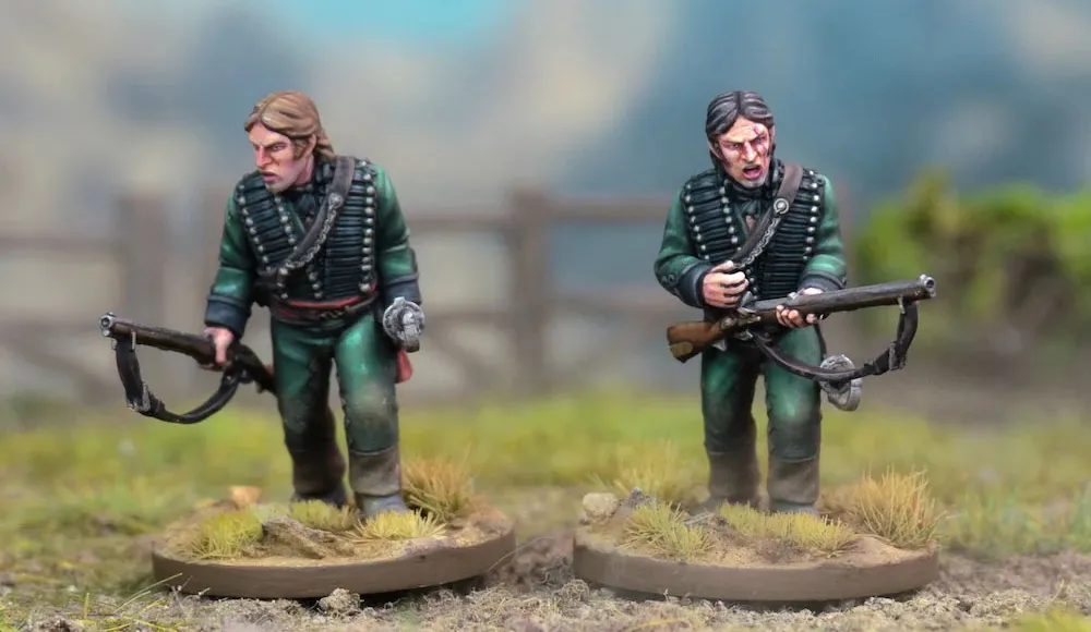 Two detailed miniatures of soldiers from the Sharpe’s Rifles series are displayed on textured bases, set against a blurred natural background. Both figures wear green uniforms with black crossbelts; one holds a rifle and has a serious expression, while the other appears to be in a more contemplative pose. The bases feature tufts of grass, enhancing the realistic terrain effect.