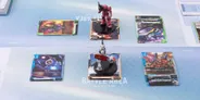 A tabletop scene features two Gundam miniatures positioned on black bases, one in red and white and the other in pink. Surrounding them are several colorful cards displaying various Gundam characters and abilities, with some cards featuring vibrant artwork of mobile suits in action. The play area has a light blue geometric background, enhancing the futuristic theme of the Gundam Card Game.