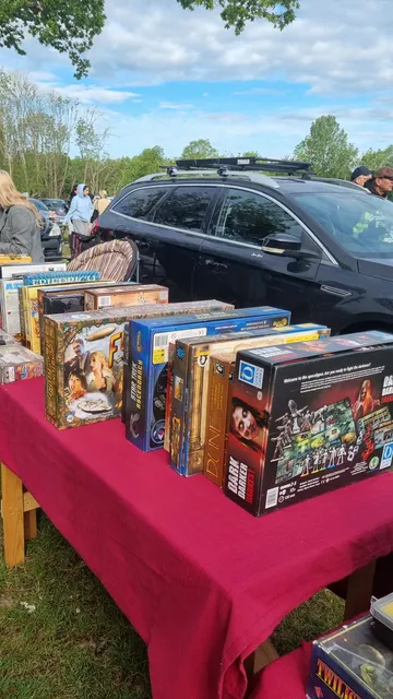 Outdoor car boot sale with red tablecloths displaying board games, DVDs, and books. People browsing and parked cars in background.
