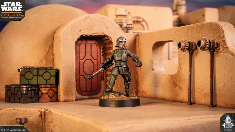 A detailed miniature of a character stands in front of a rounded, tan structure resembling a building from the Star Wars universe, with a red door and various mechanical elements visible. The character is dressed in a green uniform, holding a weapon, and is positioned on a rocky base. Nearby, there are several colorful crates in green, orange, and black, adding to the scene's immersive atmosphere.