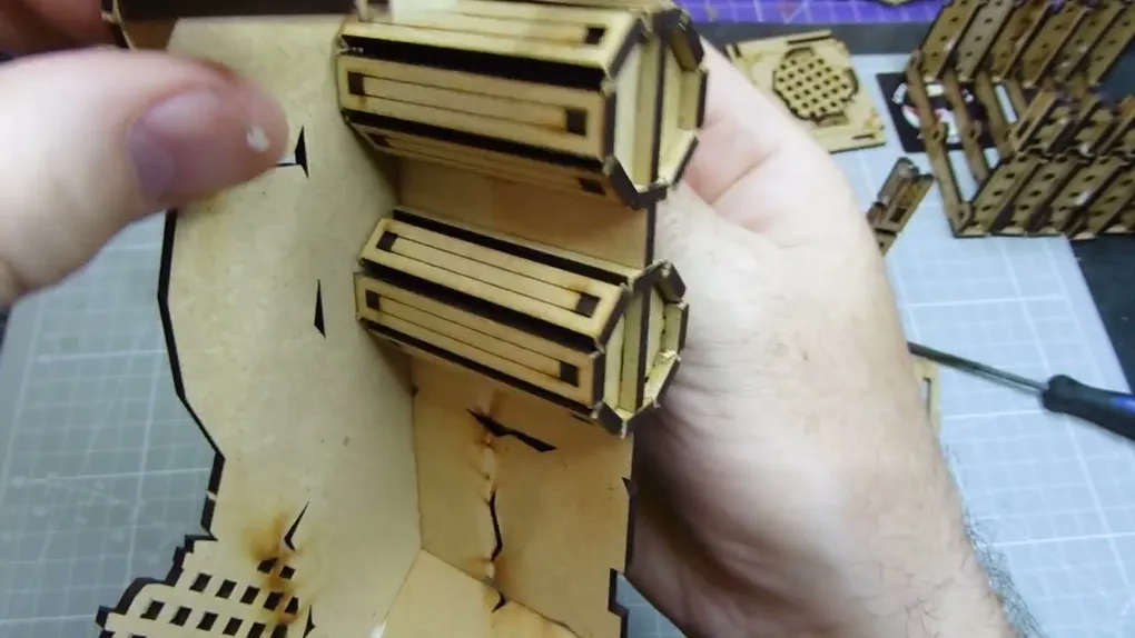 A close-up view of a partially assembled Orc Mega Bunker model from TT Combat, specifically the Orc Sektor section. The image shows a hand holding a piece of the model, which features intricate cut-out designs and layered components. The background includes a grid pattern, suggesting a crafting or modeling workspace.