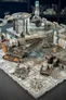 A detailed modular sci-fi terrain setup features a large, industrial-looking vehicle with tracks and a raised platform, surrounded by various structures and pipes. In the scene, two miniature figures are positioned on the terrain, one near a stack of crates and the other on a walkway. The ground is textured with cracked tiles and a circular grate, enhancing the gritty, futuristic atmosphere of the setting.