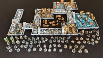 A detailed 15mm scale dungeon setup features modular tiles arranged to create a labyrinthine environment, complete with stone walls and various furniture pieces like crates and barrels. Scattered throughout the dungeon are numerous painted miniatures representing a brave group of heroes and a variety of monsters, including orcs and giant spiders. The scene is set against a dark background, enhancing the vibrant colors of the miniatures and the intricate details of the dungeon tiles.