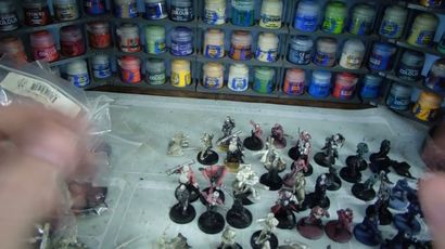 A variety of Heresy Miniatures models are arranged on a work surface, showcasing a mix of painted and unpainted figures in various poses. In the background, shelves filled with paint pots in a range of colors, including blues, greens, and reds, provide a vibrant backdrop. The models include soldiers and fantastical characters, some with detailed bases, while the surface beneath them is marked with paint splatters and tools of the hobby.