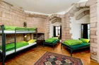 A cozy room in a medieval castle features two sets of black metal bunk beds with bright green bedding, providing a comfortable sleeping arrangement. The walls are made of light-colored stone, and there are arched stone columns that add to the historical ambiance. A patterned area rug covers the wooden floor, and large windows allow natural light to fill the space, enhancing the atmospheric setting.