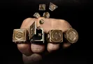 Four fantasy-themed rings are displayed on a hand, each featuring intricate designs and a hidden compartment for miniature RPG dice. The rings, crafted from brass with an aged finish, include motifs like a skull and various runes. Several polyhedral dice are shown emerging from one of the rings, highlighting their unique function as wearable dice vaults.