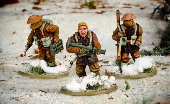 Three multi-part British and Canadian winter infantry figures are depicted in a snowy landscape. The central figure, with short blond hair, is holding a rifle and wearing a brown coat, while the other two figures are dressed in winter gear, one carrying a set of explosives and the other holding a sidearm. The bases of the figures are adorned with patches of snow and tufts of grass, enhancing the wintery scene.