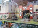 A detailed miniature display features a vibrant, stylized entrance with a skull motif and colorful tribal patterns, set against a textured wall. In the foreground, two miniatures are positioned: one is a green-skinned character with a red shoulder pad, and the other is a darker figure, both appearing ready for action. Surrounding them are various terrain elements, including rocks, a tire, and a wooden crate, enhancing the scene's immersive quality.