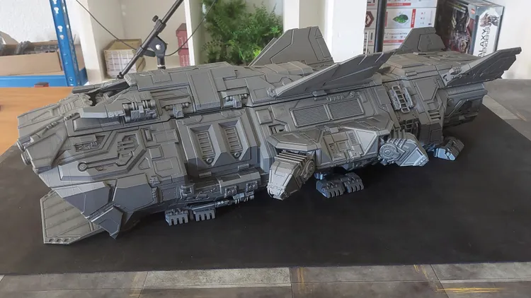 A large, intricately designed spaceship model in a gray color, featuring angular shapes and mechanical details. The model has prominent wings and various textures, showcasing its futuristic design. It is positioned on a dark tabletop, with some greenery visible in the background.