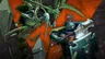 A Space Marine in dark green armor stands prominently in the foreground, wielding a large, glowing hammer and a shield adorned with intricate text and skull motifs. To the left, a Necron warrior, characterized by its skeletal frame and glowing green accents, raises a scythe-like weapon. The background features a dynamic orange geometric pattern, enhancing the tension of the scene.