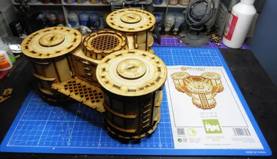 A detailed model of the Sector 3 Silo Hub from TT Combat is prominently displayed on a blue cutting mat. The structure features multiple cylindrical silos with intricate wooden textures and circular tops, showcasing a combination of open latticework and solid panels. In the background, various paint pots and a few other miniature models are visible, along with the instruction sheet for the Silo Hub, which includes an illustration of the assembled model.